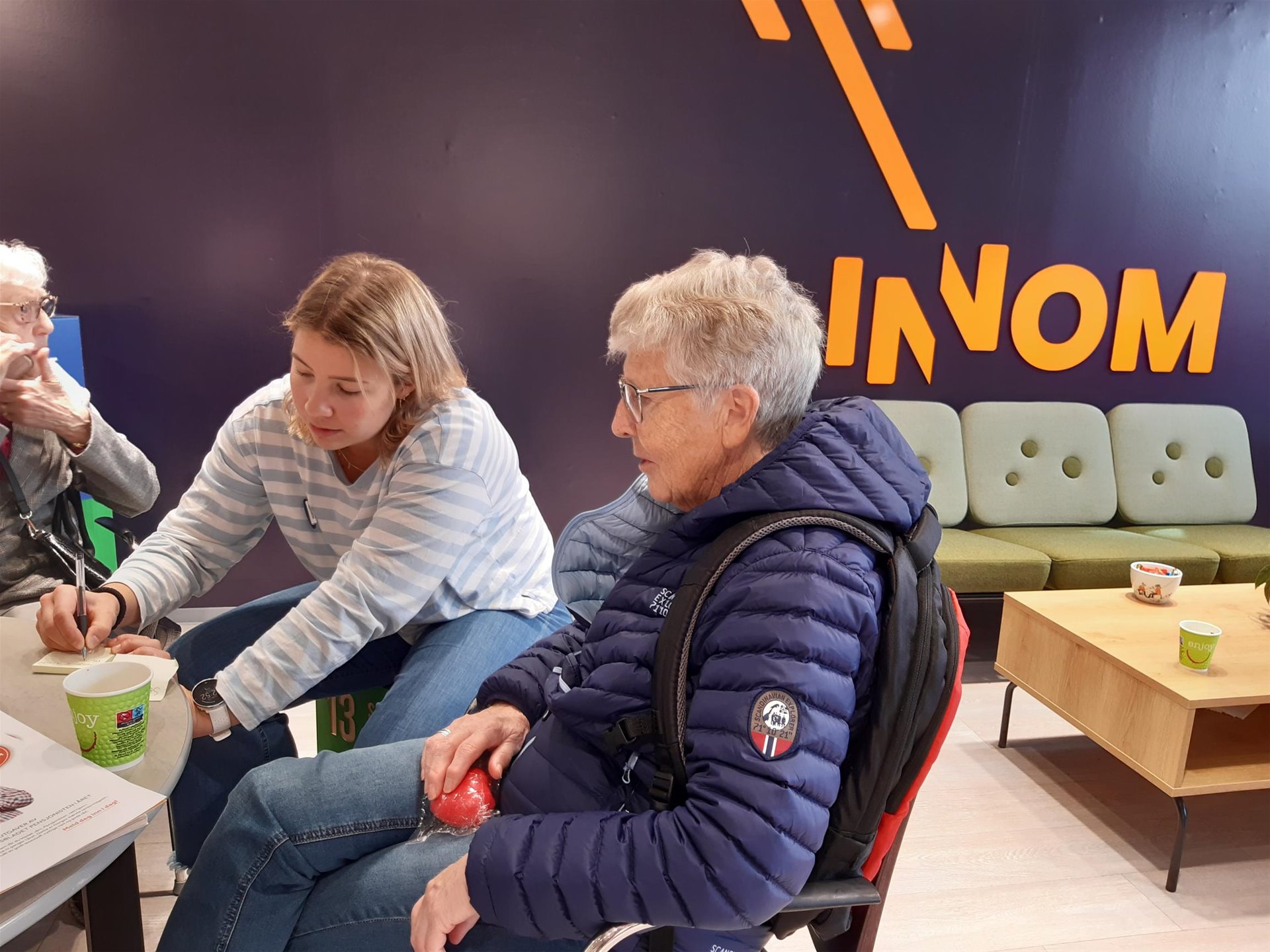Ung og eldre kvinne skriver ned et forslag på en lapp. Det står Innom på en lilla vegg. Bilde.